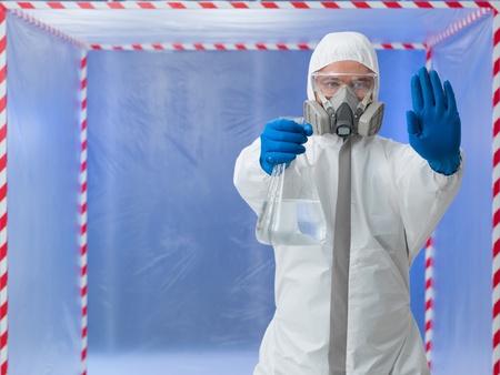 person in biohazard protective suit wearing a breathing mask calling a halt by raising one hand while holding a bottle of transparent colorless liquid in the other standing in front of a containment tentの写真素材
