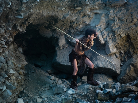 caucasian woman dressed as a caveman holding a spear, getting ready for hunting, close to a rocky...