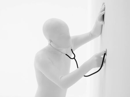 faceless man dressed in white with stethoscope in his ears, listening to a wall with hands on it の写真素材