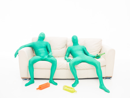 Two green men faceless, tired, sitting on a couch with bottles of drink in hand on white backgroundの写真素材