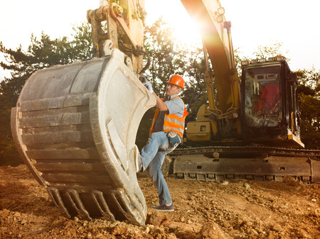 male engineer standing next to excavator on construction site outdoors, with sunlight in background