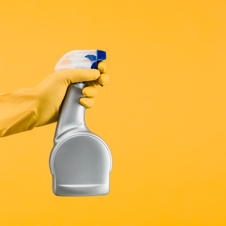 hand with rubber glove holding cleaning spray container on yellow background