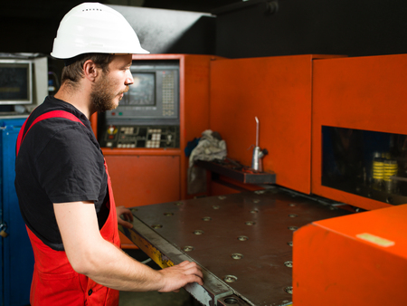 right side view of a worker in red overalls, wearing a white protective helmet, standing, looking at...