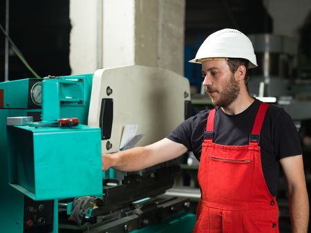 left side view of a worker, standing, wearing red overalls, and a white protective helmet, operating the control panel of an industrial bending machine, painted in turquoise and white, with another machine in the background, in an industrial settingの写真素材