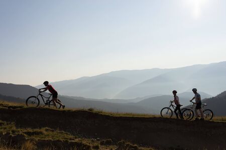bikers on the mountain pushing their bikes uphill at sunsetの写真素材
