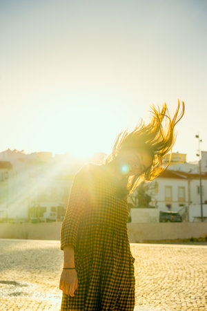 Portrait of a girl in a city square during a sunny day, looking at the camera and laughing against the sky with sun rays filtering through her  windy hairの写真素材