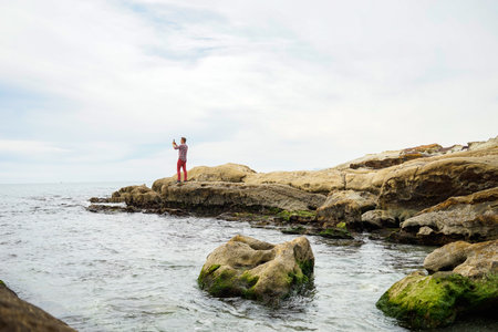 Traveler man taking photo with smartphone from the ocean rocky shore, foggy day