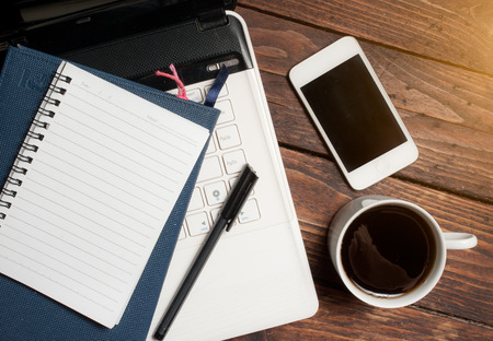Office desk with laptop computer, notebook, mobile smartphone and coffee cup on wood,morning lightの写真素材