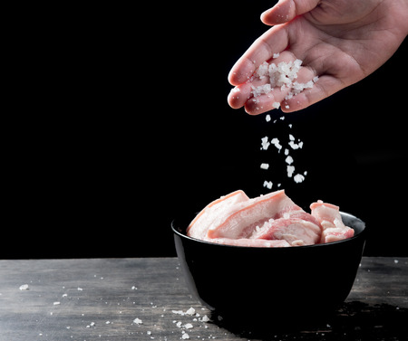 Hand sprinkling salt on raw meat of pork over black background.の写真素材