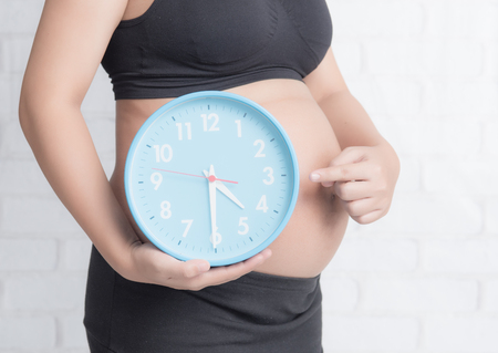 pregnant woman holding a big round clock against white brick wallの写真素材