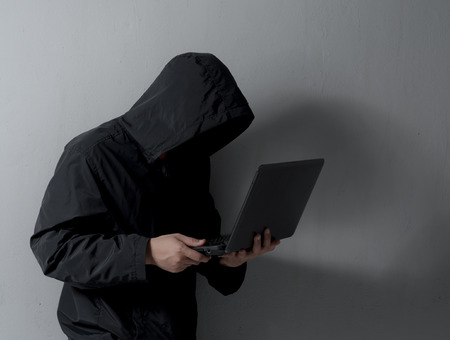 Hacker with hidden face in a hoodie using a laptop against white brick wall