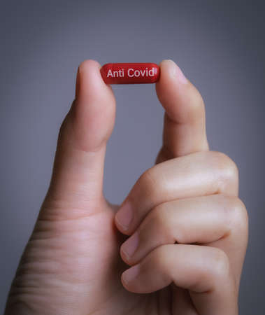Male hand holding a medical red capsule pill between fingers against gray background,Coronavirus,Covid-19 Cure,Anti-Covid 19,anti flu drug pillの写真素材