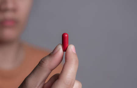 Female hand holding a medical red capsule pill between fingers against gray backgroundの写真素材