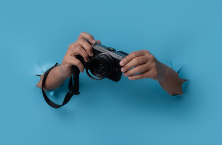 Female hand holding camera breaks through blue paper background.の写真素材
