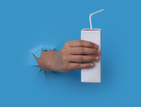Female hand with blank packet carton juice & milk breaks through blue paper background.の写真素材