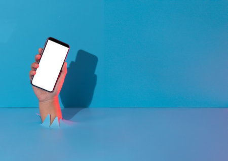 Hand holding smartphone with white empty screen showing device,breaks through blue paper background.の写真素材