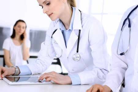 Group of doctors at work in hospital with patient in a queue at the background. Physician filling up medical documents or prescription while standing at reception desk. Data in medicine and health care conceptの写真素材