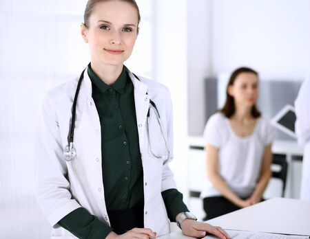 Doctor woman at work with patient and colleague at background. Physician filling up medical documents or prescription while standing in hospital reception desk. Data in medicine and health care conceptの写真素材