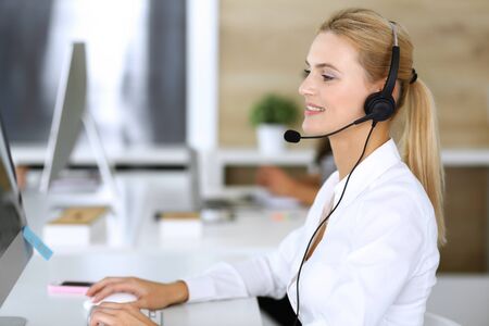 Blonde business woman using headset for communication and consulting people at customer service office. Call center. Group of operators at work at the background