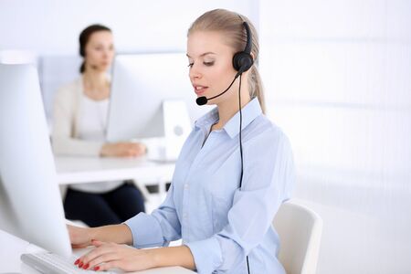 Call center office. Beautiful blonde woman using computer and headset for consulting clients online. Group of operators working as customer service occupation. Business people conceptの写真素材