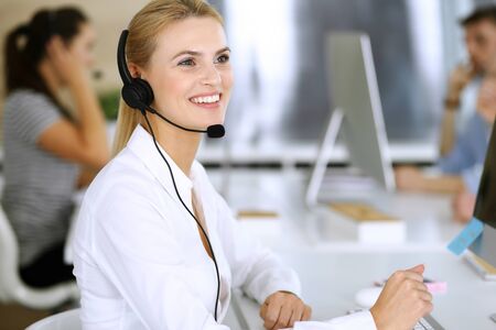 Blonde business woman using headset for communication and consulting people at customer service office. Call center. Group of operators at work at the backgroundの写真素材