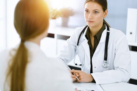 Woman-doctor and patient sitting and talking at sunny hospital office. Green color blouse suits to therapist. Medicine conceptの写真素材