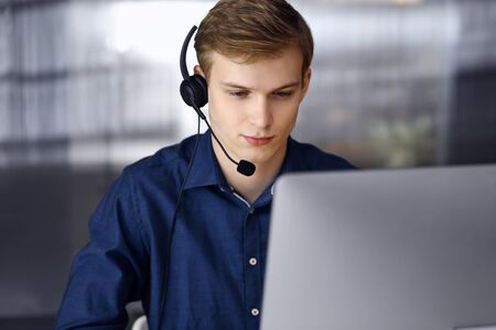 Young blond businessman using headset and computer at work. Startup business means working hard and...
