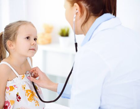 Doctor examining a child by stethoscope in sunny clinic. Happy smiling girl patient dressed in bright color dress is at usual medical inspectionの写真素材