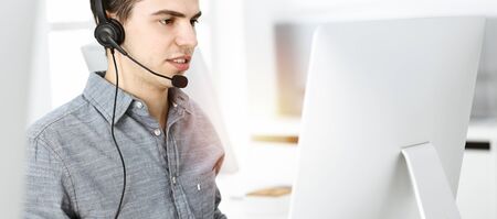 Casual dressed young man using headset and computer while talking with customers online in sunny...