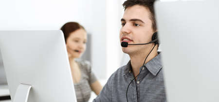 Casual dressed young man using headset and computer while talking with customers online. Group of...
