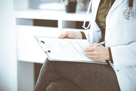 Unknown woman-doctor writing something at clipboard while sitting at the chair, close-up. Therapist wearing green blouse at work is filling up medication history record. Medicine conceptの写真素材