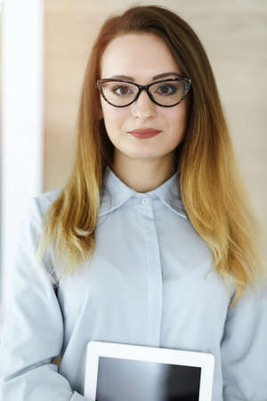 Business woman headshot in sunny office. Unknown businesswoman standing straight with tablet computer. Young accountant or secretary looks goodの写真素材