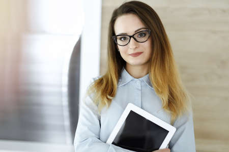 Business woman headshot in sunny office. Unknown businesswoman standing straight with tablet computer. Young accountant or secretary looks goodの写真素材