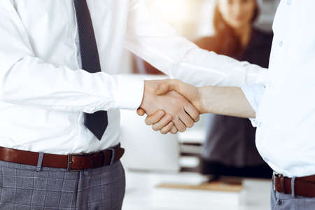 Two businessmen shaking hands in sunny office, close-up. Happy and excited business woman stands with raising hands at the background. Business people conceptの写真素材