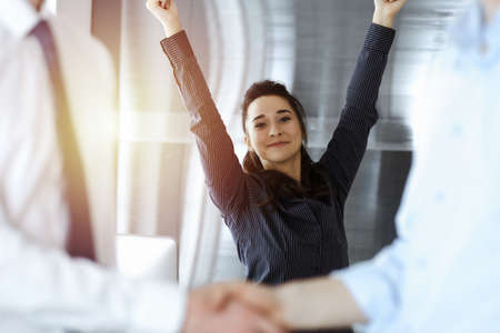 Happy and excited business woman raising hands because of success agreement at meeting. Two men...