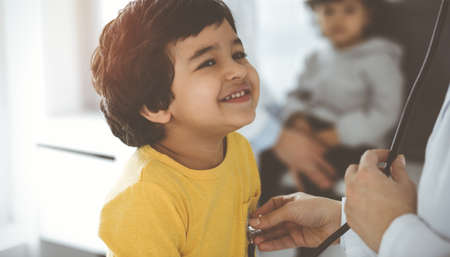 Woman-doctor examining a child patient by stethoscope in sunny clinik. Cute arab boy at physician appointmentの写真素材