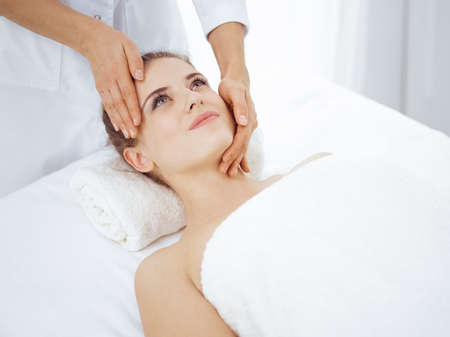 Young and blonde woman enjoying facial massage in spa salon. Beauty concept