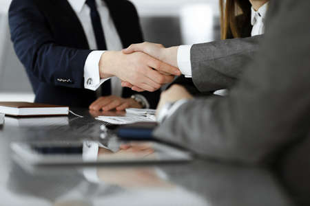 Unknown businessman shaking hands with his colleague or partner above the glass desk in modern office, close-up. Business people group at meetingの写真素材
