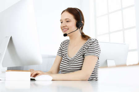 Casual dressed young woman using headset and computer while talking with customers online. Call center, business conceptの写真素材