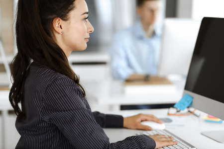 Business woman using computer at workplace in modern office. Brunette secretary or female lawyer smiling and looks happy. Working for pleasure and successの写真素材