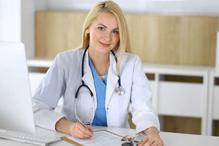 Doctor woman at work while sitting at the desk in hospital or clinic. Blonde cheerful physician filling up medical records form. Data and best service in medicine and healthcareの写真素材