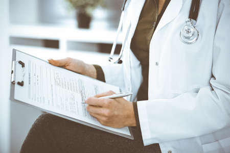 Unknown woman-doctor writing something at clipboard while sitting at the chair, close-up. Therapist wearing green blouse at work is filling up medication history record. Medicine conceptの写真素材