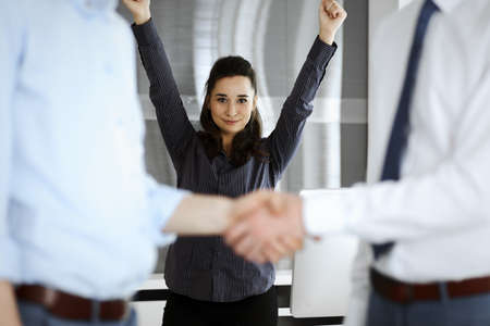 Happy and excited business woman raising hands because of success agreement at meeting. Two men...