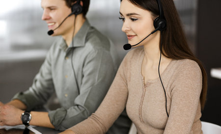 Two young people in headsets are talking to the clients, while sitting at the desk in an office. Focus on woman. Call center operators at workの写真素材