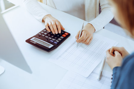 Accountant checking financial statement or counting by calculator income for tax form, hands close-up. Business woman sitting and working with colleague at the desk in office. Audit conceptの写真素材