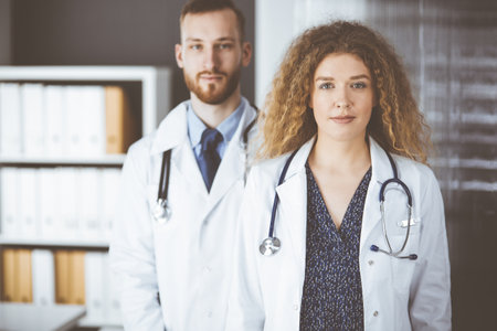 Red-bearded doctor with female colleague discussing current disease therapy while sitting at working place in clinic. Team work in medicineの写真素材