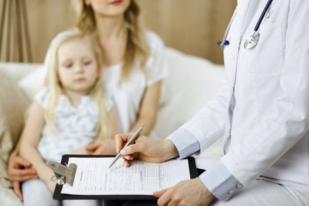 Doctor and patient. Pediatrician using clipboard while examining little girl with her mother at home. Sick and unhappy child at medical exam. Medicine conceptの写真素材