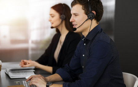 Call center operators at work. Two young people in headsets are talking to the clients, while sitting in sunny officeの写真素材