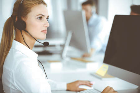 Blonde business woman using headset for communication and consulting people at customer service office. Call center. Group of operators at workの写真素材
