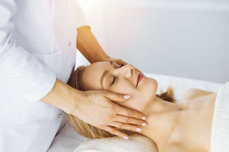 Beautiful caucasian woman enjoying facial massage with closed eyes in sunny spa salon. Relaxing treatment in medicineの写真素材